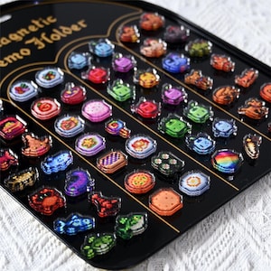May include: A black magnetic memo holder filled with colourful pixelated magnets. The magnets feature various designs, including food items, geometric shapes, and abstract patterns. The text "magnetic memo holder" is visible on the holder.