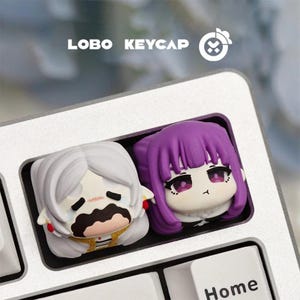 May include: Two custom keycaps with anime-style character designs. One keycap has white hair and a sad expression, the other has purple hair and a neutral expression. The keycaps are set on a silver keyboard. The text "LOBO KEYCAP" is visible.
