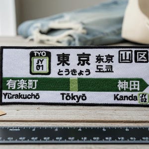 Może przedstawiać: Prostokątna naszywka z japońskim tekstem i transliteracjami angielskimi. Naszywka zawiera słowa "Tōkyō", "Yūrakuchō" i "Kanda" w kolorze zielonym i białym. Czarna linijka i pusta etykieta znajdują się na pierwszym planie.