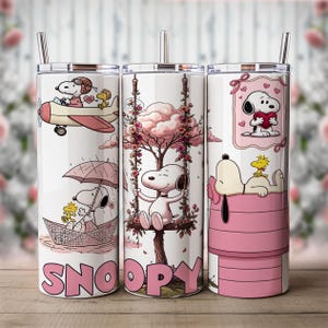 May include: Three white cylindrical tumblers with metal straws featuring Snoopy and Woodstock illustrations. Designs include Snoopy flying a plane, sitting on a floral swing, and resting on a pink doghouse. The word SNOOPY is printed in pink.