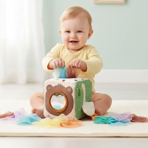 May include: A soft fabric sensory cube toy featuring a brown bear face with a circular mirror, a green side with a white goose illustration, and colorful sheer fabric squares that can be pulled out from the top opening of the cube.