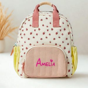 Könnte beinhalten: Ein cremefarbener Rucksack mit Erdbeermuster. Der Rucksack hat einen rosa Griff und Träger sowie eine rosa Vordertasche mit dem Namen "Amelia" in Schreibschrift. Gelbe Taschen befinden sich an den Seiten.