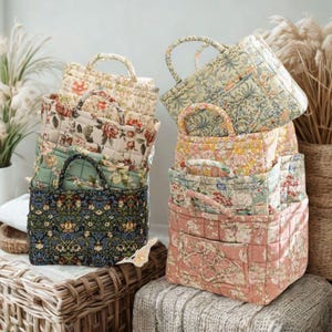 May include: A stack of quilted tote bags with floral patterns in various colors. The bags have sturdy handles and are made of fabric with designs including roses, leaves, and other botanical elements. The bags are stacked on a wicker basket and a knitted blanket.