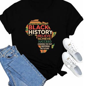 Pode incluir: Camiseta preta com um mapa colorido da África e as palavras "Black History" e frases inspiradoras. A imagem também mostra jeans azuis e tênis brancos, criando um look casual e estiloso.