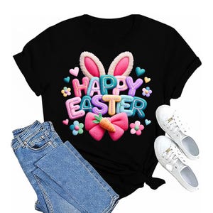 Peut inclure: T-shirt noir avec un motif coloré "Joyeuses Pâques" représentant des oreilles de lapin, des fleurs et un nœud avec une carotte. L'image comprend également un jean bleu et des baskets blanches.