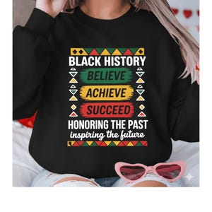 Pode incluir: Camisola preta com o texto "BLACK HISTORY BELIEVE ACHIEVE SUCCEED HONORING THE PAST inspiring the future" em branco, verde, amarelo e vermelho. O design inclui padrões geométricos em preto, amarelo, verde e vermelho.