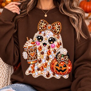 Peut inclure: Un sweat-shirt marron à col rond orné d'un motif de fantôme décoré de fausses pierres précieuses et de perles. Le fantôme porte un nœud à motif léopard, tient un gobelet à café léopard avec de la crème fouettée et un seau citrouille.