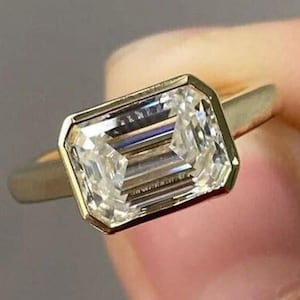 May include: A gold ring featuring a large, rectangular, clear gemstone. The gemstone is set in a bezel setting, and the band is a simple, polished gold. The ring is held up against a neutral background.