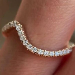 Puede incluir: Un anillo de banda de oro curvado engastado con pequeñas piedras preciosas redondas y transparentes. El anillo se muestra en un dedo, con la banda curvándose hacia arriba en el centro. Las piedras preciosas están muy juntas, creando un brillo continuo.