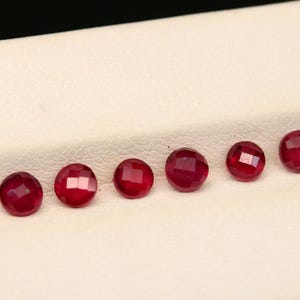 May include: A row of faceted, round, deep red gemstones. The gems are set against a white background, with each stone reflecting light. The gems are likely rubies, and are arranged in a line.