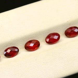 May include: A collection of faceted, oval-shaped, deep red gemstones arranged on a cream-colored surface. The gems have a brilliant cut, reflecting light. The background is a neutral cream color.