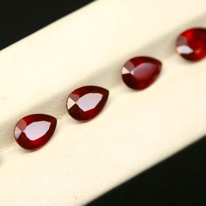 May include: A collection of six pear-shaped, faceted, deep red gemstones. The gems are arranged in a diagonal line on a cream-colored surface, with a black background. The gemstones have a high luster and reflect light.