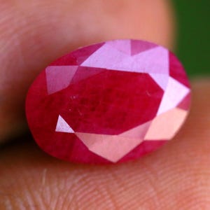 Puede incluir: Una piedra preciosa de rubí de forma ovalada y facetada con un color rojo intenso y vibrante. La piedra presenta una superficie pulida con múltiples facetas geométricas que reflejan la luz, creando reflejos y sombras distintos en su cuerpo.