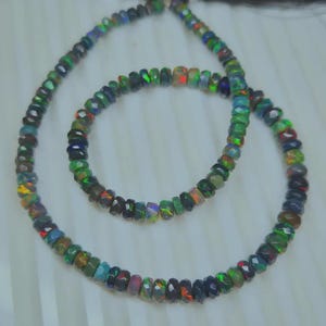 May include: Two strands of faceted opal beads in a necklace and bracelet set. The beads display a range of colors, including green, blue, red, and black. The necklace has a black tassel.