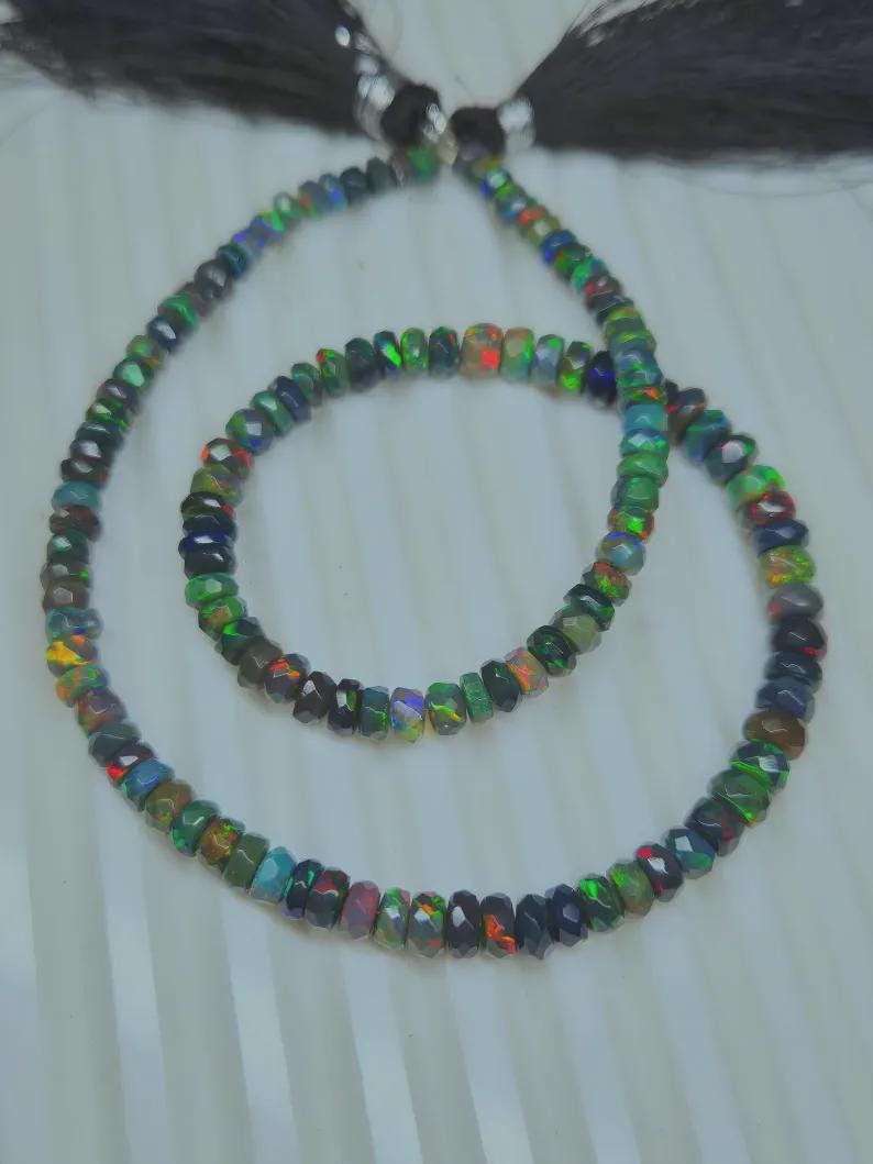 May include: Two strands of faceted opal beads in a necklace and bracelet set. The beads display a range of colors, including green, blue, red, and black. The necklace has a black tassel.