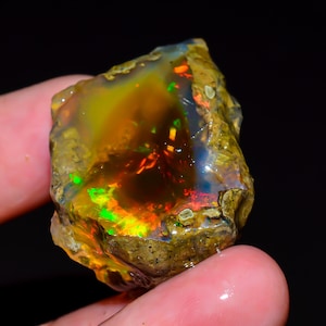 May include: A rough opal gemstone with a vibrant display of colors. The stone features flashes of green, orange, and red within a translucent, golden-brown matrix. The opal is held in a hand, showcasing its natural, unpolished texture.