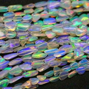 Op de afbeelding: Close-up van strengen iriserende opaal kralen. De kralen zijn een mix van heldere en opalescente kleuren, met flitsen van blauw, groen en geel. De kralen zijn glad en gepolijst en lijken aan elkaar geregen te zijn.