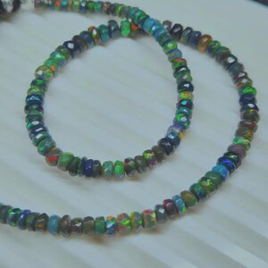 May include: A necklace composed of faceted, iridescent opal beads. The beads display a range of colors, including green, blue, red, and black. The necklace is displayed on a white surface.