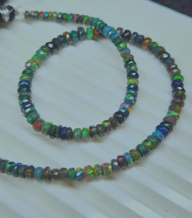 May include: A necklace composed of faceted, iridescent opal beads. The beads display a range of colors, including green, blue, red, and black. The necklace is displayed on a white surface.