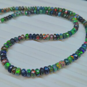 May include: A necklace composed of small, faceted beads in shades of green, blue, red, and black. The beads are strung together to form a circular shape. The necklace is displayed on a white surface.