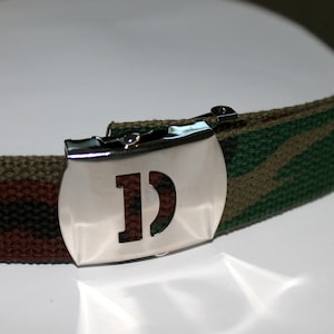 May include: A camouflage patterned fabric belt with a silver buckle featuring a cut-out letter "D".