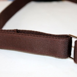 Puede incluir: Una correa ajustable marrón con herrajes metálicos plateados. La correa está hecha de tejido tejido y tiene un acabado liso.