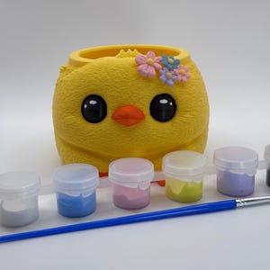 May include: A yellow chick-shaped paint-your-own kit with large black eyes, an orange beak, and a floral crown. Includes six paint pots in various colours and a blue paintbrush. A fun craft project for children.