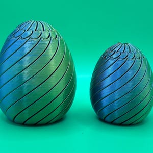May include: Two decorative egg-shaped objects with a gradient color scheme of blue and green. The eggs have a spiral design with carved lines and a textured top. The eggs are on a green background.
