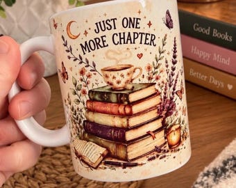 Op de afbeelding: Een witte keramische mok met een stapel kleurrijke boeken, een dampende theekop, vlinders en een maansikkel. De tekst "JUST ONE MORE CHAPTER" staat in zwarte letters. Het ontwerp bevat bloemmotieven, een klein open boek en een kaars.