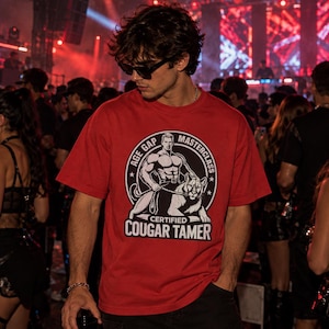 Pode incluir: Uma t-shirt vermelha de manga curta com um gráfico circular a preto e branco de uma figura musculada a segurar uma trela presa a um puma. O texto diz AGE GAP MASTERCLASS e CERTIFIED COUGAR TAMER em letras maiúsculas brancas.