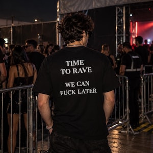 Puede incluir: Una camiseta negra de manga corta con texto blanco en la espalda que dice TIME TO RAVE WE CAN FUCK LATER. La prenda es usada por una persona vista desde atrás, con una tela de apariencia lisa y opaca en un entorno nocturno al aire libre.