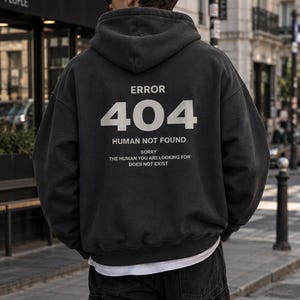 Könnte beinhalten: Ein anthrazitfarbener Kapuzenpullover mit weißem Text auf der Rückseite, der ERROR 404 HUMAN NOT FOUND SORRY THE HUMAN YOU ARE LOOKING FOR DOES NOT EXIST lautet. Der Hoodie hat lange Ärmel, eine Kapuze und einen sichtbaren weißen Saum.