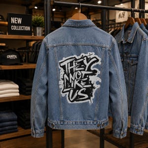 Puede incluir: Una chaqueta de mezclilla azul con un gráfico grande de estilo grafiti en blanco y negro en la espalda con el texto "THEY NOT LIKE US" en una fuente audaz y estilizada. La chaqueta tiene cuello clásico, mangas largas y costuras visibles.