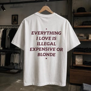 May include: A white short-sleeved t-shirt featuring dark red text on the back that reads: EVERYTHING I LOVE IS ILLEGAL EXPENSIVE OR BLONDE. The shirt is displayed on a wooden hanger, showcasing a crew neckline and a standard fit design.