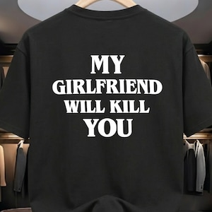 May include: Black t-shirt with the white text "MY GIRLFRIEND WILL KILL YOU" in a bold, sans-serif font. The shirt is hanging on a wooden hanger, with a blurred background of a closet.