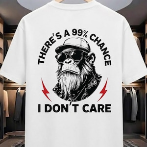 Könnte beinhalten: Weißes T-Shirt mit einer Schwarz-Weiß-Illustration eines Schimpansen mit Sonnenbrille und Lederjacke. Der Text lautet "THERE'S A 99% CHANCE I DON'T CARE" in Schwarz, mit roten Blitz-Akzenten.