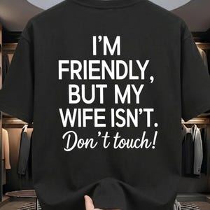 May include: Black t-shirt with the white text "I'M FRIENDLY, BUT MY WIFE ISN'T. Don't touch!" The shirt has short sleeves and a crew neck. The text is in a bold, sans-serif font.
