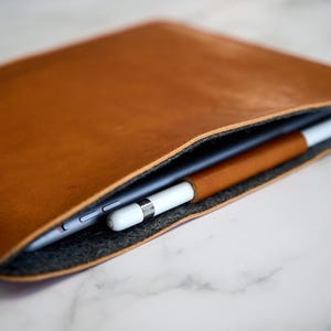 Può includere: Una custodia per tablet in pelle marrone con un porta stilo abbinato. La custodia è aperta, rivelando un tablet e uno stilo bianco. L'interno è foderato in feltro grigio. La pelle ha una consistenza liscia.