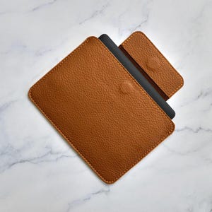 Può includere: Una custodia per tablet in pelle marrone con un lembo di chiusura magnetica abbinato. La custodia ha una superficie strutturata e un oggetto nero a contrasto è parzialmente visibile all'interno. Le cuciture sono di colore marrone chiaro.