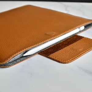 Può includere: Una custodia in pelle color cuoio con superficie testurizzata e rivestimento interno in feltro grigio. La custodia presenta una patta con chiusura magnetica e contiene una penna stilo bianca inserita nell'apertura sul bordo superiore.