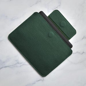 Può includere: Una custodia in pelle verde scuro con una superficie strutturata. La custodia rettangolare ha angoli arrotondati e una piccola chiusura a bottone circolare. Un oggetto nero è parzialmente visibile all'interno, insieme a un accessorio verde più piccolo abbinato.