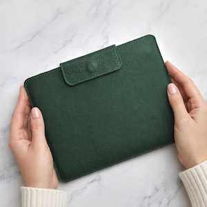 May include: A dark green leather tablet case with a flap closure. The case is rectangular with rounded corners and a textured surface. The flap has a button closure. The case is held by two hands.