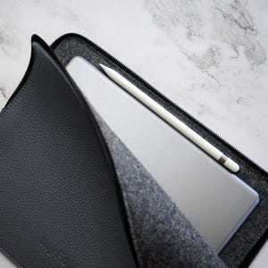 May include: A gray leather tablet case with a black zipper, holding a silver tablet and a white stylus. The case is open, revealing a gray felt interior. The stylus is resting on the tablet.