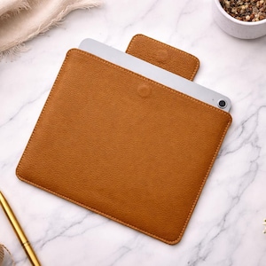 May include: A tan leather tablet sleeve with a textured surface and a flap closure. The sleeve features a rectangular shape with rounded corners and visible stitching along the edges. A silver tablet is partially inserted into the protective case.