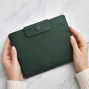May include: A dark forest green leather portfolio case with a textured pebble grain finish. The rectangular case features a flap closure secured with a matching green button snap. The edges are finished with tonal stitching along the perimeter.