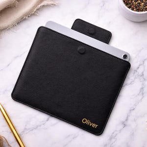 May include: A black textured leather tablet sleeve with a magnetic flap closure. The sleeve features the name Oliver embossed in gold lettering on the bottom right corner. A silver tablet is partially inserted into the black protective case.
