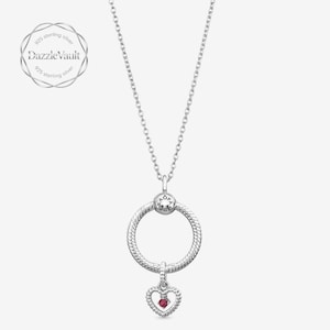Puede incluir: Un collar de plata de ley con una cadena fina y un colgante circular con un patrón de cuerda texturizado. Un dije más pequeño en forma de corazón con una piedra roja está suspendido de la parte inferior del círculo. El logo DazzleVault es visible.