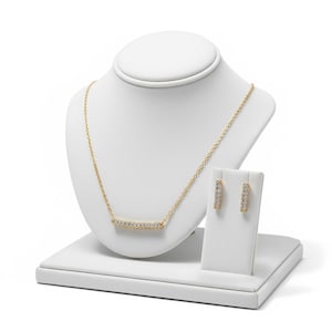 May include: A gold-colored necklace and matching earrings set displayed on a white jewelry bust and stand. The necklace features a horizontal bar pendant adorned with small, clear gemstones. The earrings are also gold-colored, with a similar gemstone design.