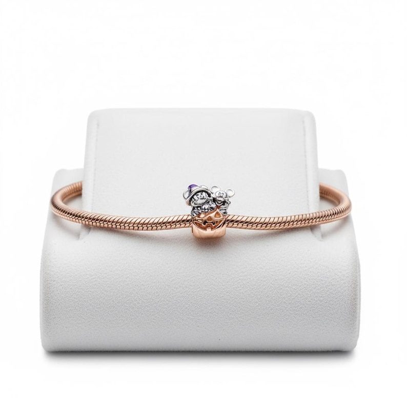 May include: A rose gold bracelet with a charm featuring two cartoon characters in Halloween costumes. The charm has silver and gold tones with purple and orange accents. The bracelet is displayed on a white stand.