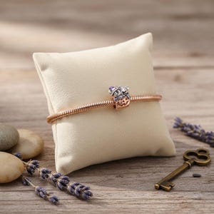 May include: A rose gold bracelet with a charm featuring a crown and purple accents, displayed on a cream-colored pillow. The bracelet is set against a wooden background with lavender sprigs and a vintage key.